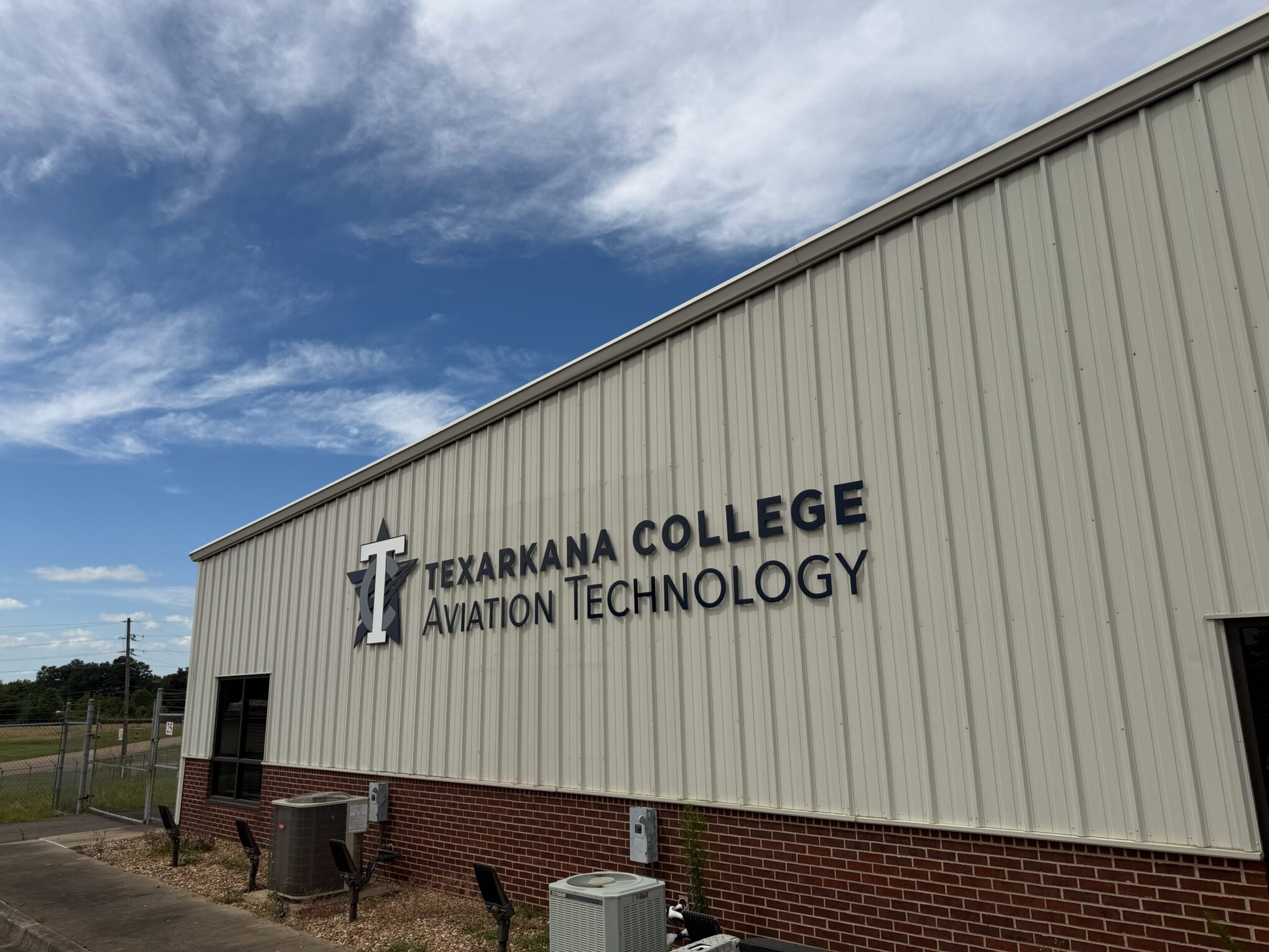 A beige and red brick building with a sign reading Texarkana College Aviation Technology under a blue sky with clouds.